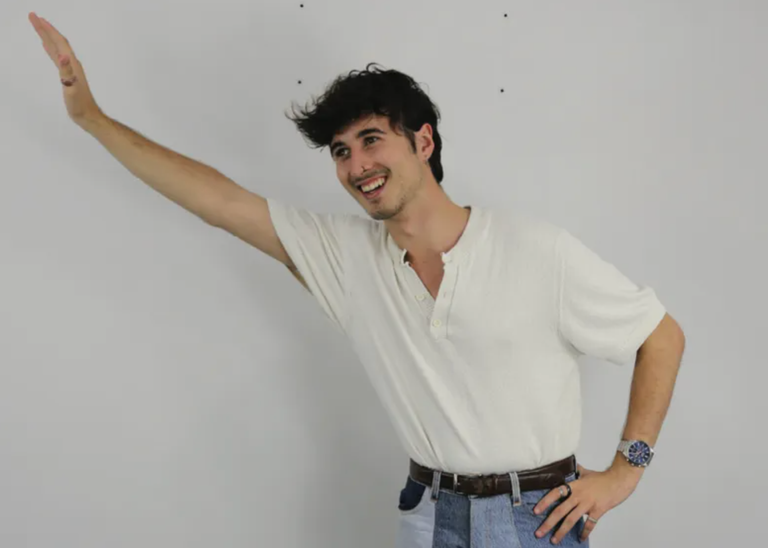 Promotional portrait of comedian Tom Holmes smiling and waving towards the side in a white shirt against a plain light wall background